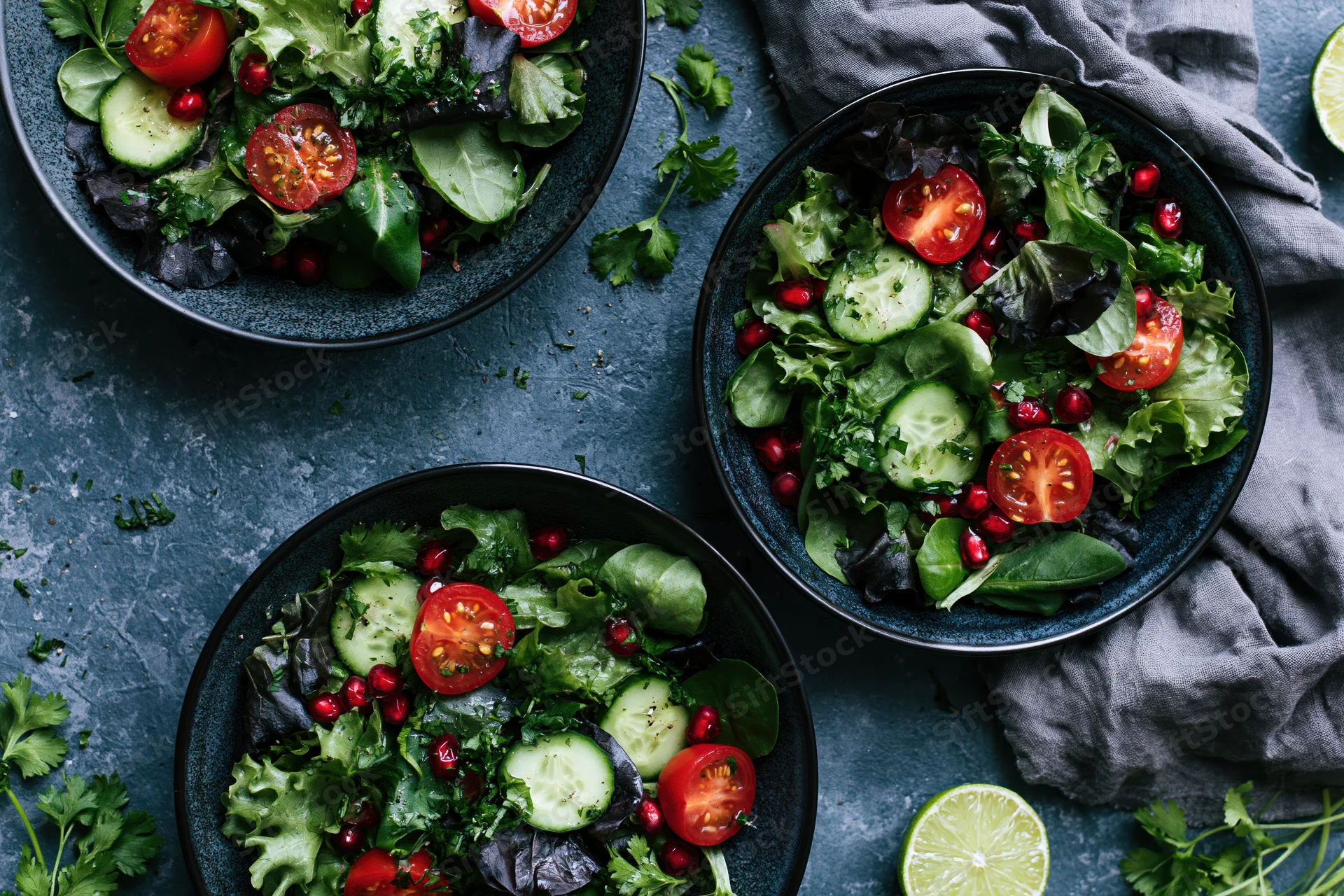 Three dark ceramic bowls filled with fresh salad topped with tomatoes cucumbers and pomegranate seeds on a dark surface