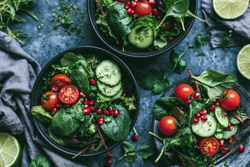 Overhead flat lay of dark ceramic bowls filled with fresh salad and garnishes on a gray surface