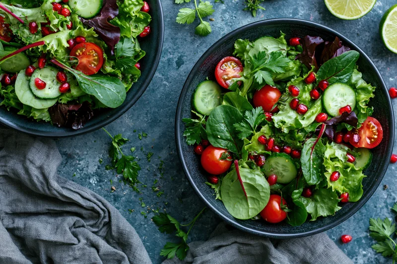 Two dark ceramic bowls filled with fresh salad of greens, cucumber, tomato, and pomegranate seeds on a gray stone surface with scattered herbs and cloth napkin