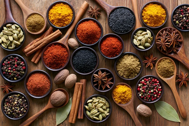 Overhead flat lay of assorted whole and ground spices arranged on wooden surface with wooden spoons and bowls