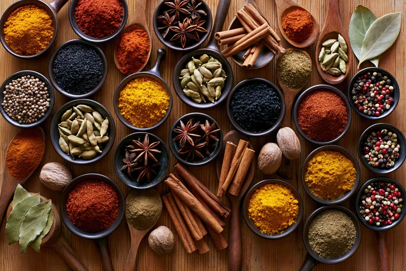 Overhead flat lay of assorted whole and ground spices arranged in bowls and spoons on wooden surface