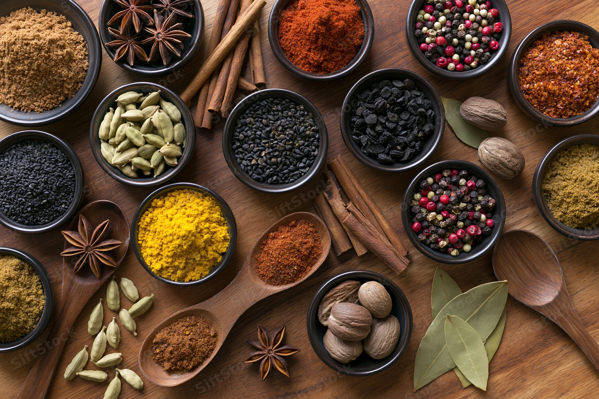 Overhead flat lay of assorted whole and ground spices arranged in bowls and spoons on wooden surface