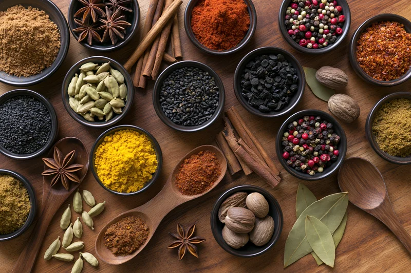 Overhead flat lay of assorted whole and ground spices arranged in bowls and spoons on wooden surface