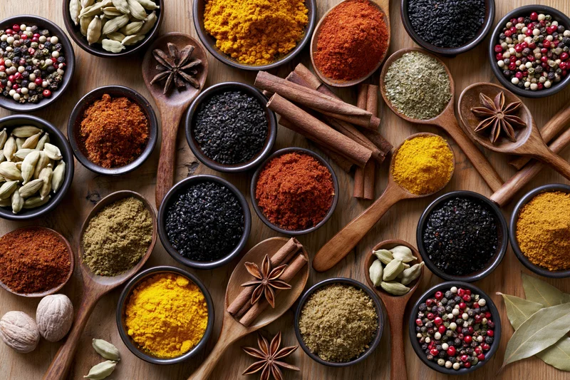 Overhead flat lay of assorted whole and ground spices arranged on wooden surface in bowls and spoons