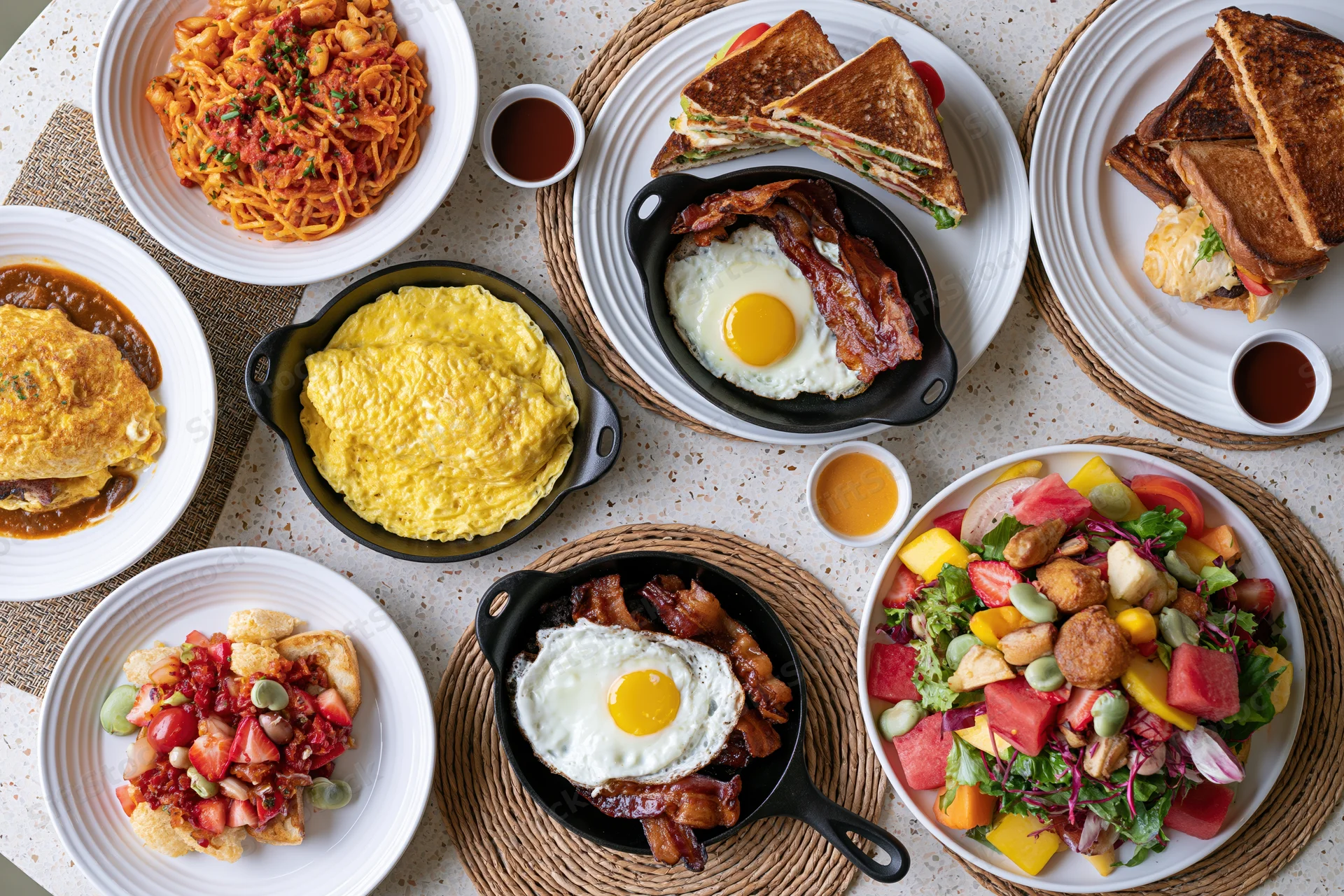 Overhead flat lay of eight diverse brunch dishes arranged on a table with plates, skillets, and woven mats