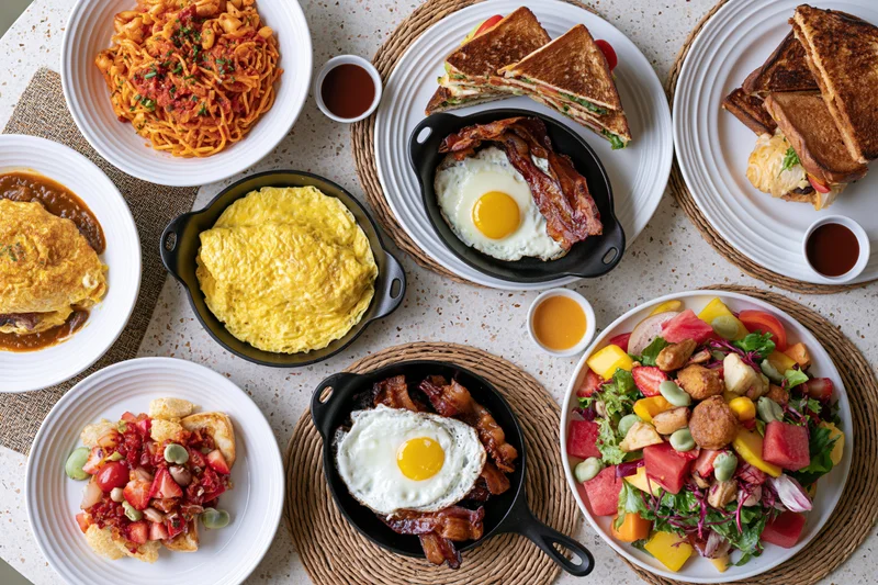 Overhead flat lay of eight diverse brunch dishes arranged on a table with plates, skillets, and woven mats