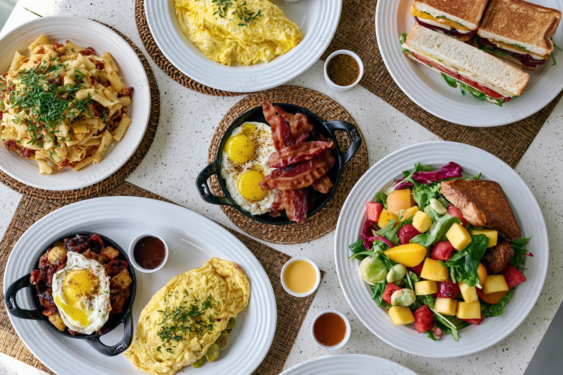 Overhead flat lay of diverse brunch dishes on a white countertop with woven placemats