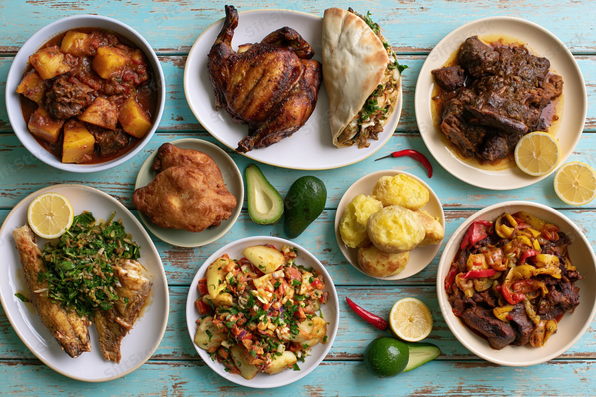 Overhead flat lay of eight varied dishes including roasted chicken, fish, beef stew, and vegetables arranged on a blue wooden table