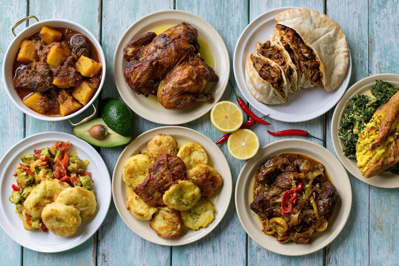 Overhead flat lay of eight dishes including roasted chicken, stews, roti, dumplings and vegetables arranged on a blue wooden table