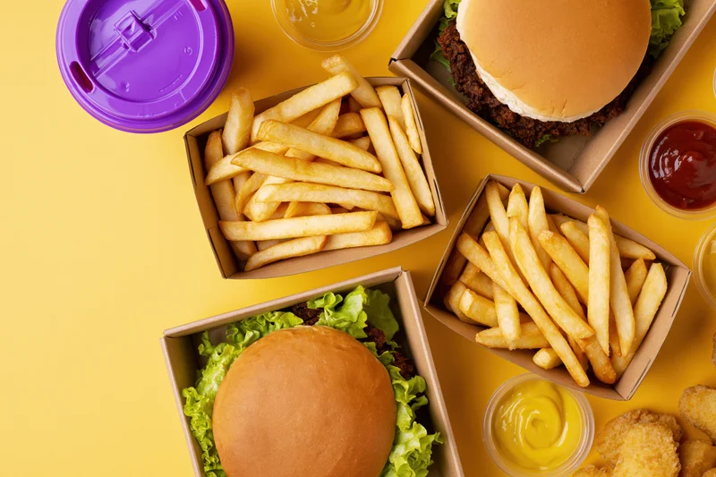 Overhead flat lay of fast food meal with two burgers, fries, sauces, and drink on yellow background