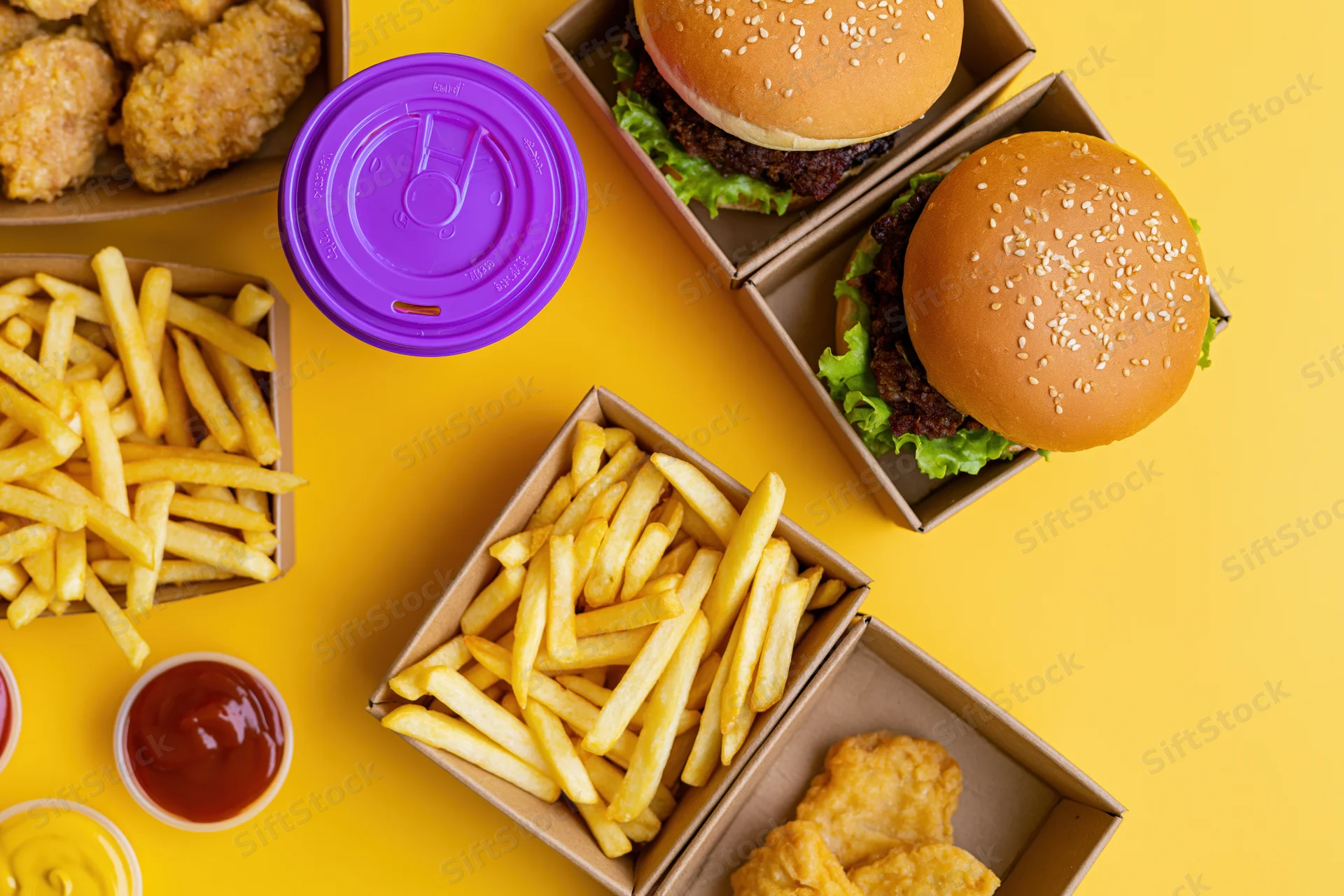 Overhead flat lay of fast food meal with burgers, fries, chicken, sauces and soda on yellow background