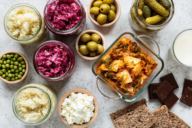 Overhead flat lay of fermented and probiotic foods arranged on a countertop including kimchi, sauerkraut, olives, pickles, yogurt, cheese, chocolate, and bread in jars and bowls