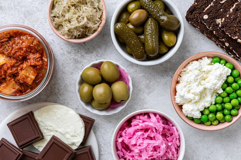 Overhead flat lay of fermented and probiotic foods arranged on a gray surface including kimchi, sauerkraut, pickles, olives, cottage cheese, peas, chocolate, yogurt, and bread in bowls and plates