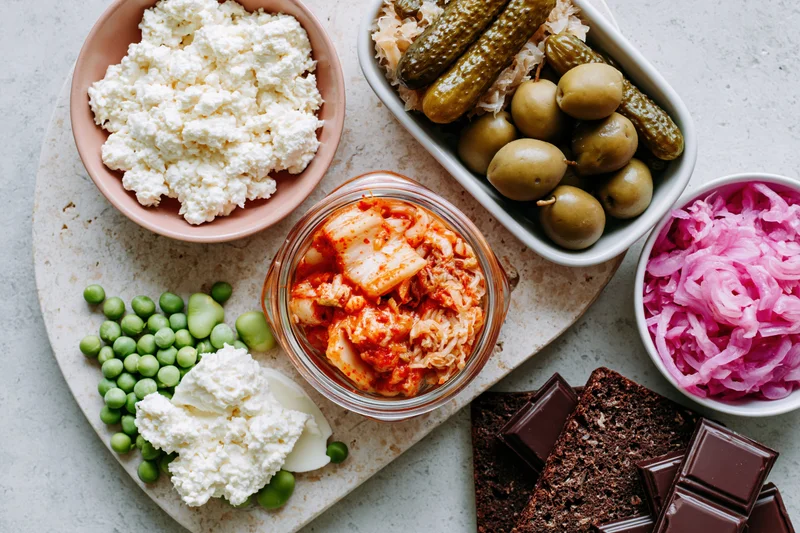Overhead flat lay of fermented and probiotic foods arranged on a tray with cottage cheese, kimchi, pickles, olives, sauerkraut, peas, chocolate, and rye bread