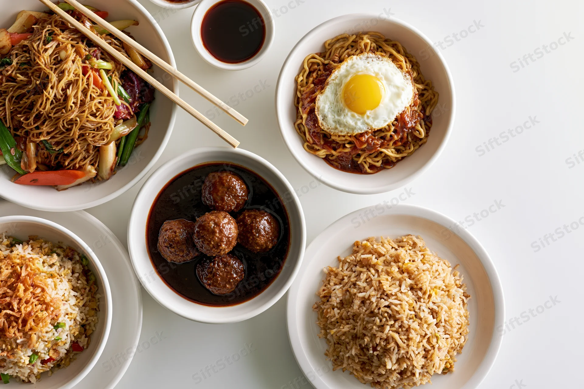 Overhead flat lay of five white ceramic bowls and plates arranged with various dishes including noodles, rice, meatballs, fried egg, and vegetables on a white surface