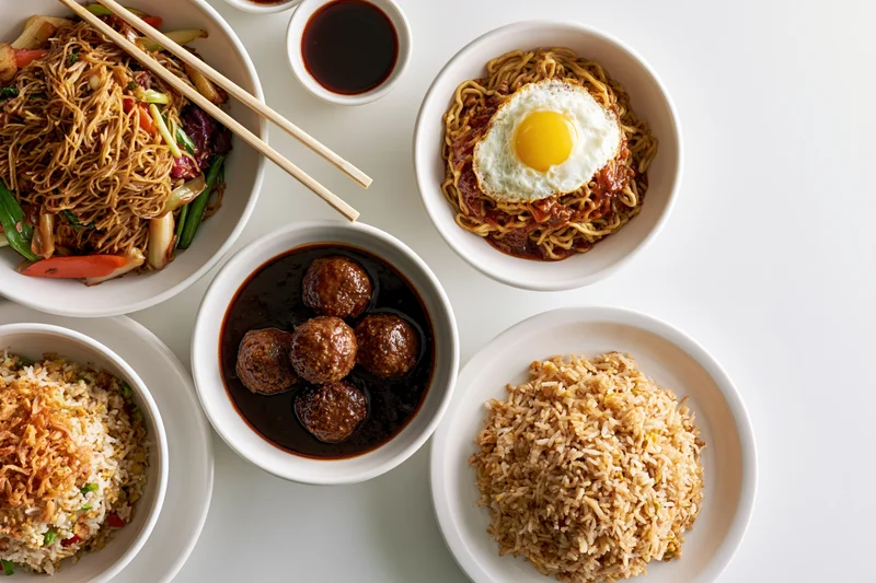 Overhead flat lay of five white ceramic bowls and plates arranged with various dishes including noodles, rice, meatballs, fried egg, and vegetables on a white surface