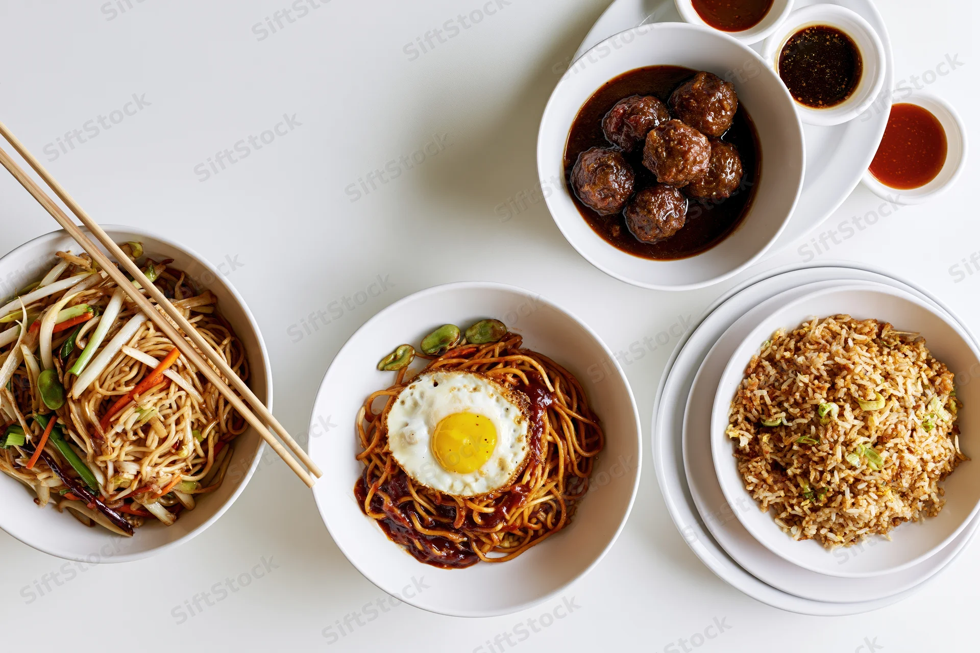 Overhead flat lay of five white ceramic bowls with noodles, fried rice, meatballs, and sauce arranged on a white surface