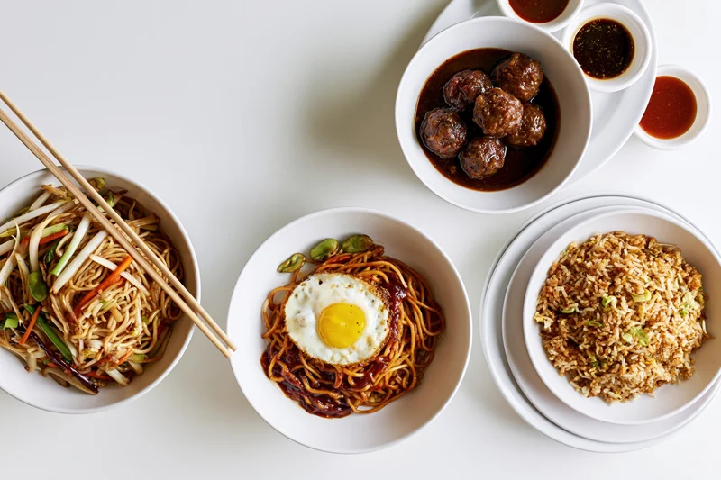Overhead flat lay of five white ceramic bowls with noodles, fried rice, meatballs, and sauce arranged on a white surface