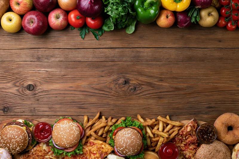 Fresh fruits and vegetables arranged at top of wooden table with fast food items at bottom