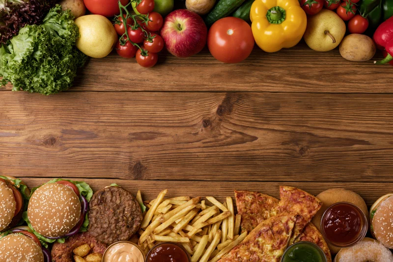 Fresh vegetables and fruits arranged at top of wooden table with fast food items at bottom