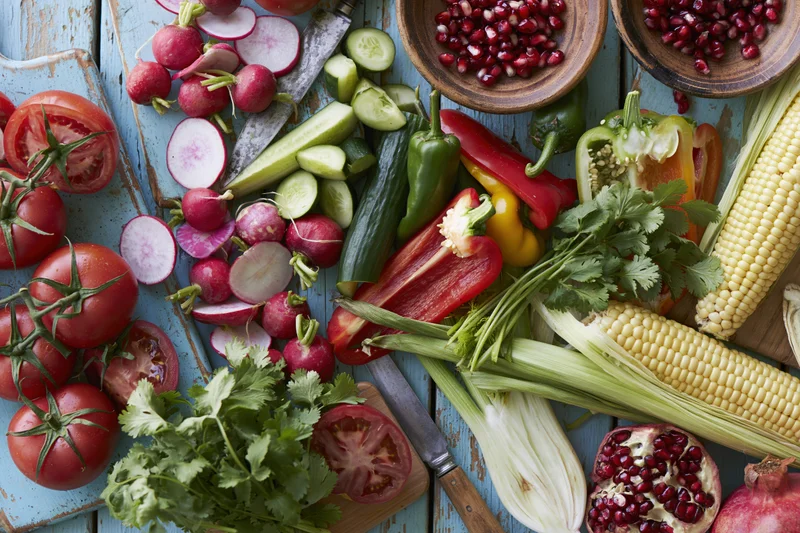 Overhead flat lay of fresh vegetables and fruits mid-prep on a blue wooden cutting board