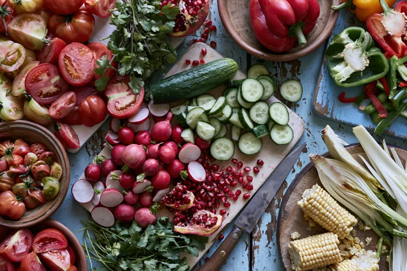 Overhead flat lay of fresh vegetables and fruits mid-prep on a wooden surface with knife and cutting board