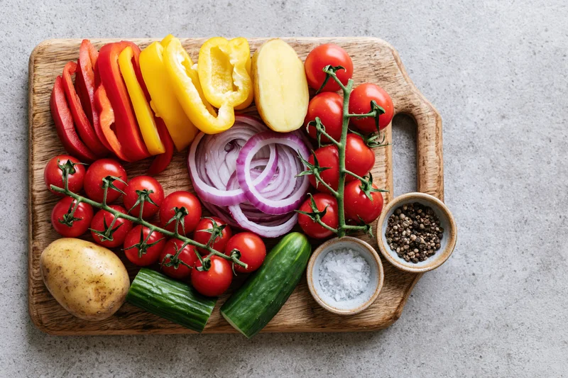 Fresh vegetables arranged on a wooden cutting board with salt and pepper bowls on a gray countertop