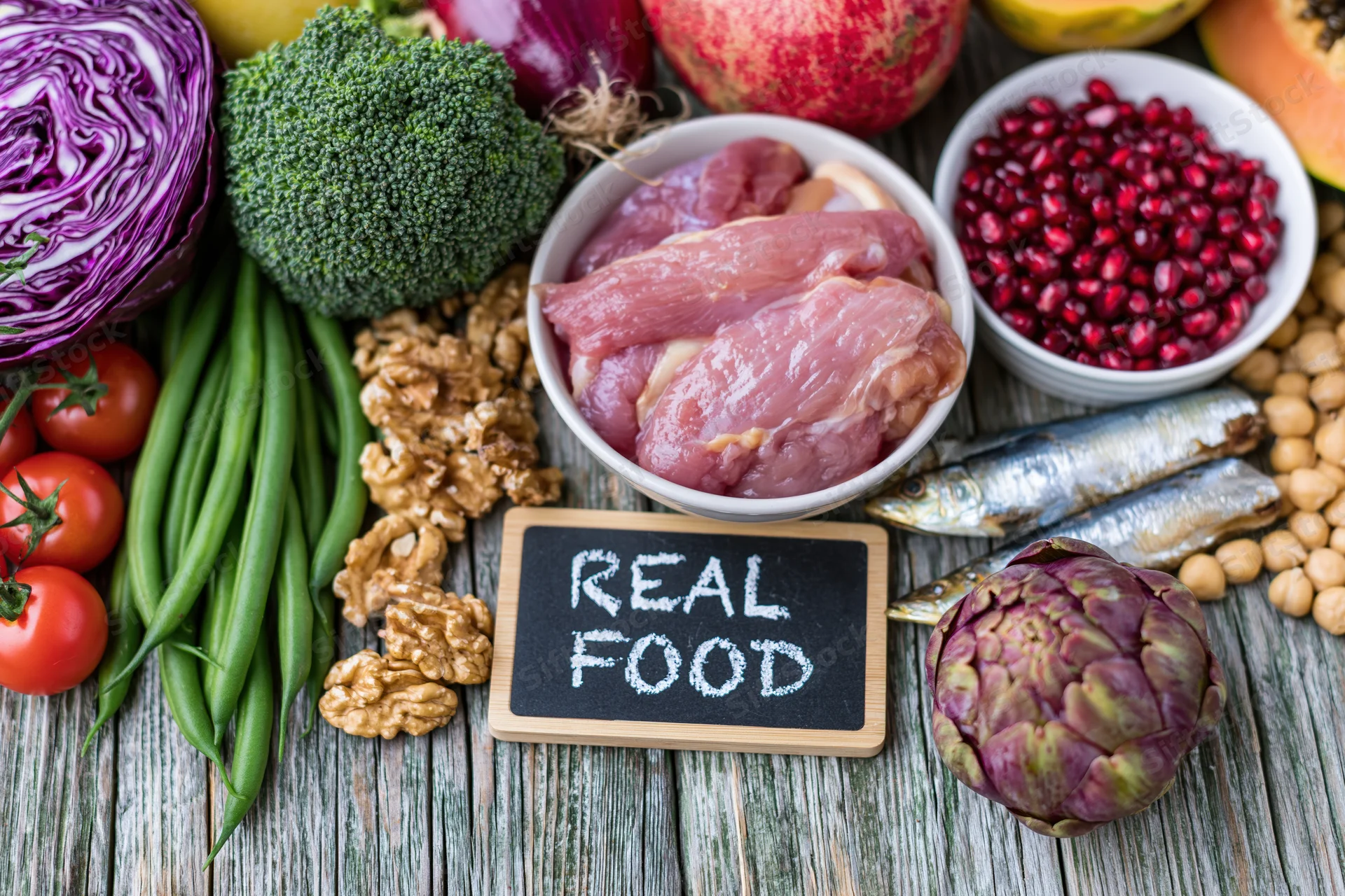 Overhead flat lay of fresh whole foods arranged around a chalkboard sign reading real food on a rustic wooden table