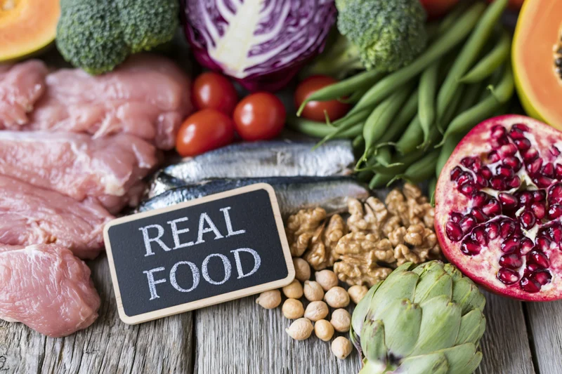 Fresh whole foods arranged around a chalkboard sign reading real food on a wooden surface