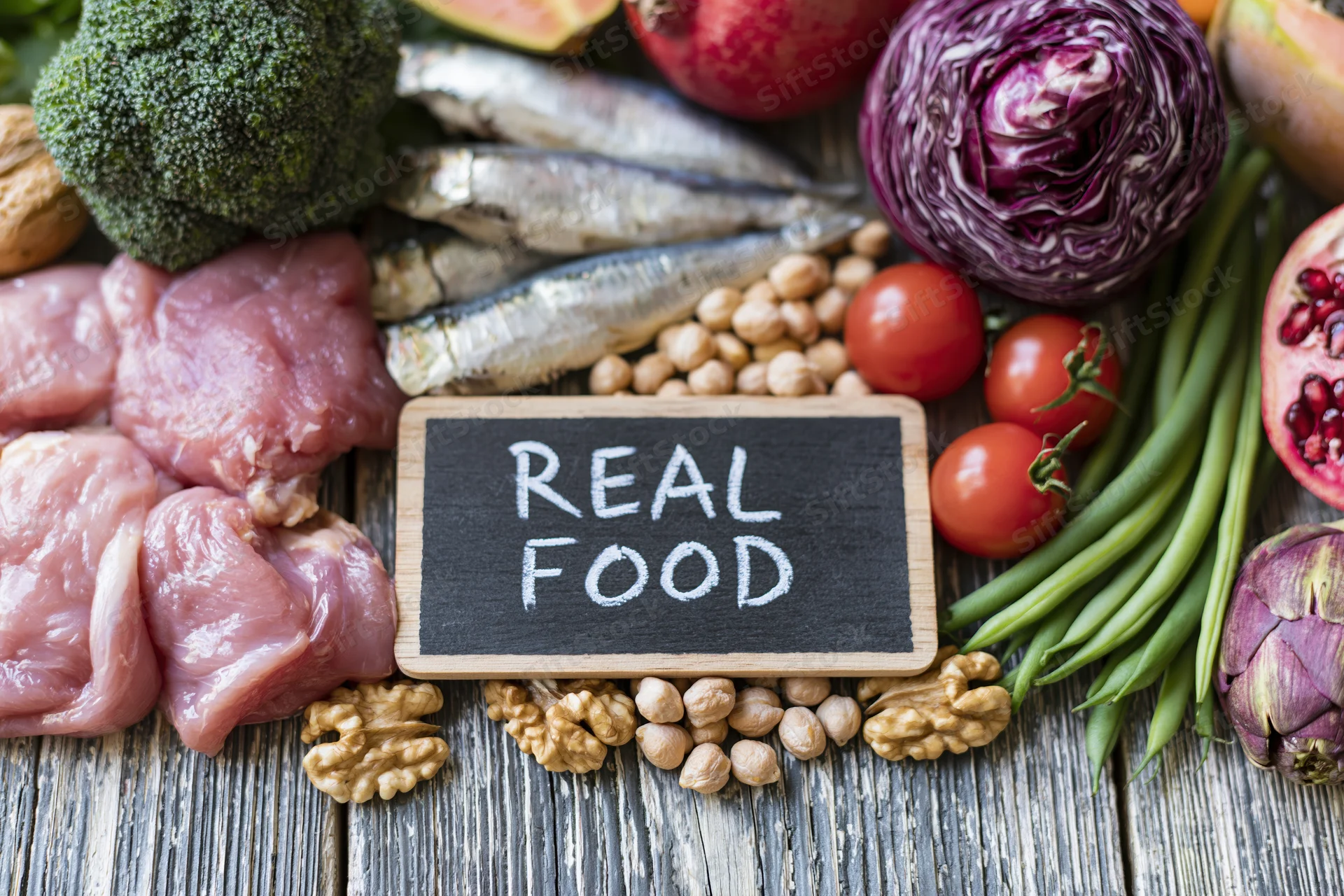 Overhead flat lay of fresh whole foods arranged around a chalkboard sign reading real food on a wooden surface
