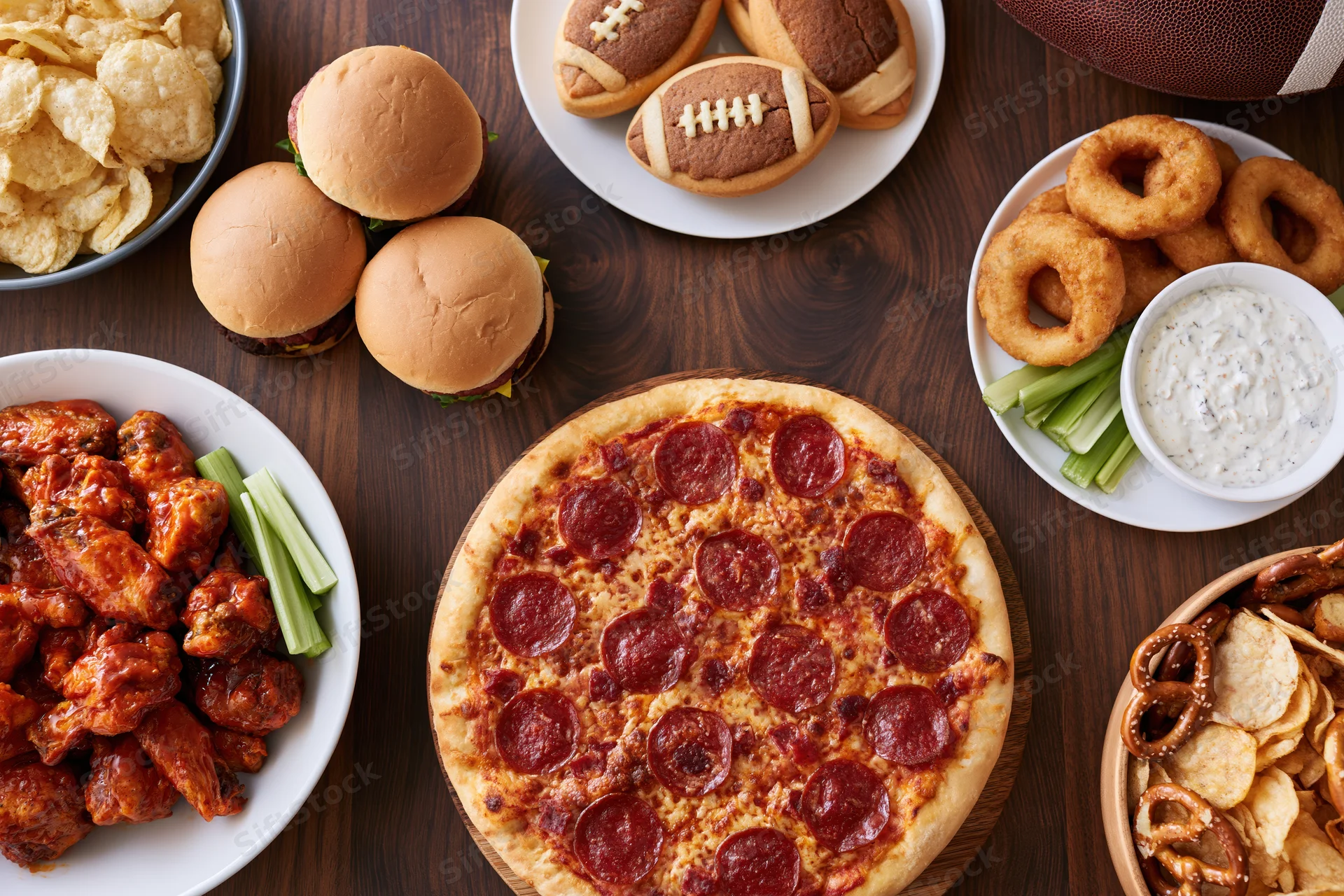 Overhead flat lay of game day party food spread on dark wood table featuring pizza, sliders, wings, onion rings, chips and football-themed snacks