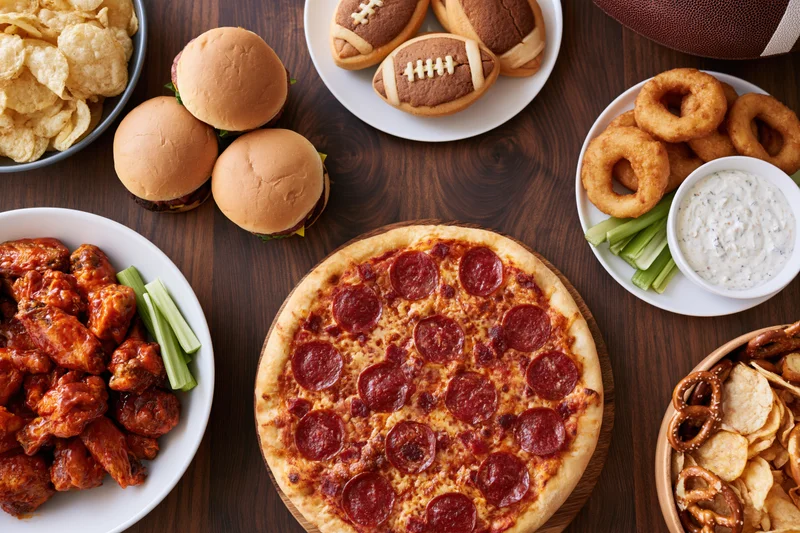 Overhead flat lay of game day party food spread on dark wood table featuring pizza, sliders, wings, onion rings, chips and football-themed snacks