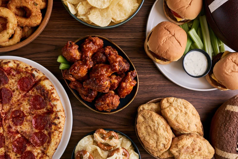 Overhead flat lay of game day party food spread on dark wooden table featuring pizza, buffalo wings, burgers, onion rings, chips, cookies, and footballs