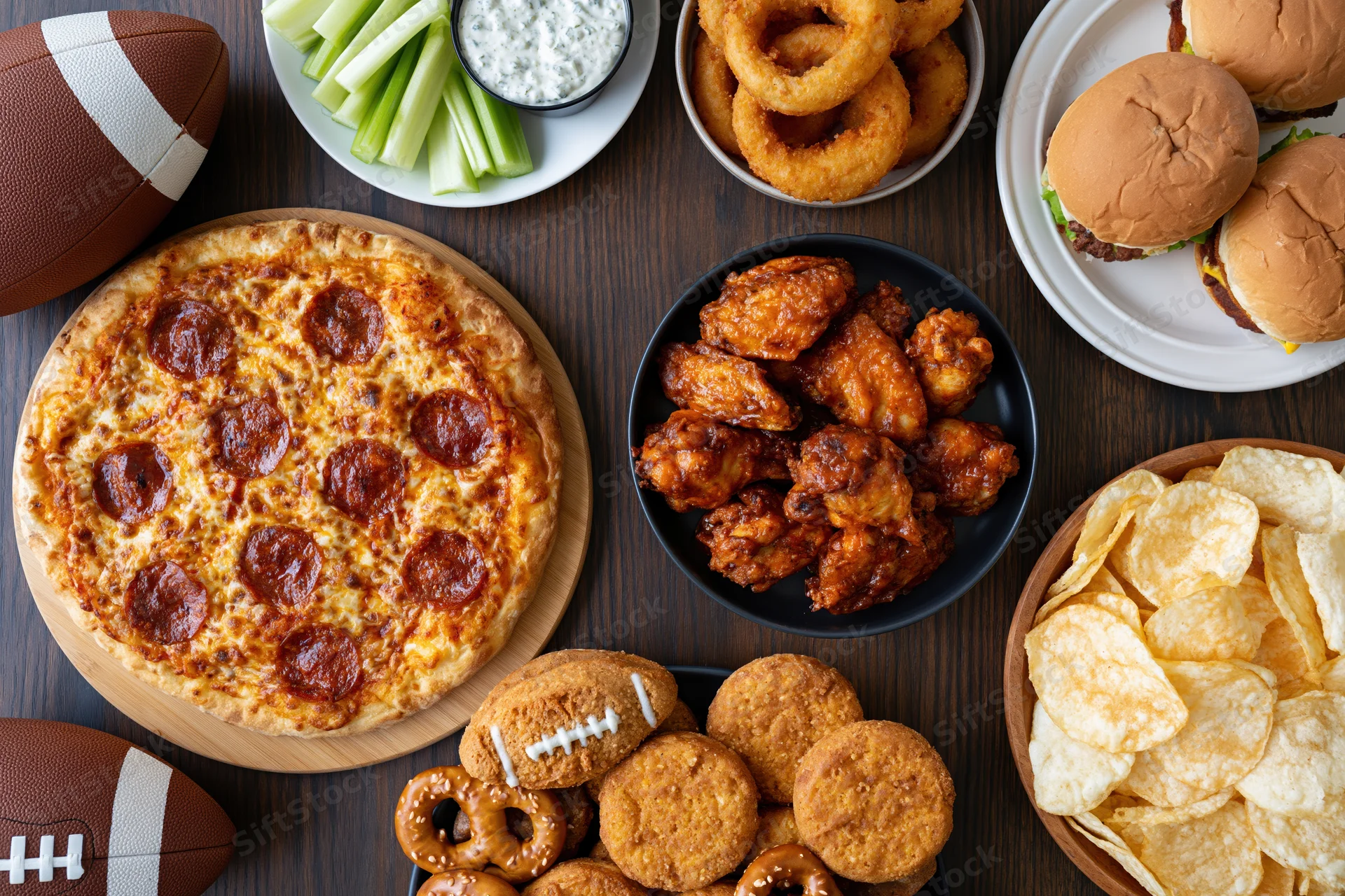 Overhead flat lay of game day party food spread on dark wood table featuring pizza, wings, burgers, chips, and football-themed snacks
