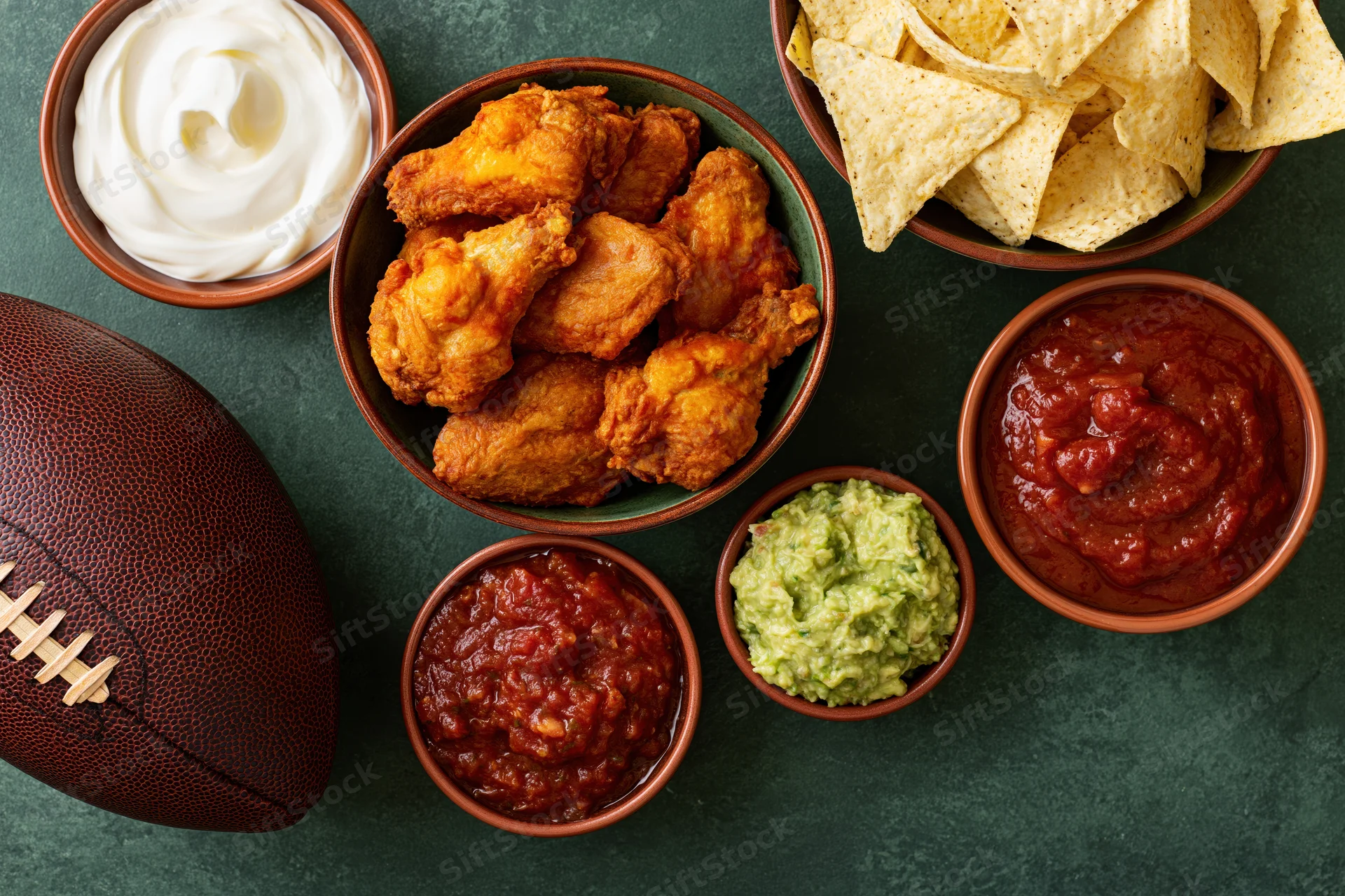 Overhead flat lay of game day snacks including buffalo chicken wings, tortilla chips, salsa, guacamole, and sour cream arranged with a football on a dark green surface