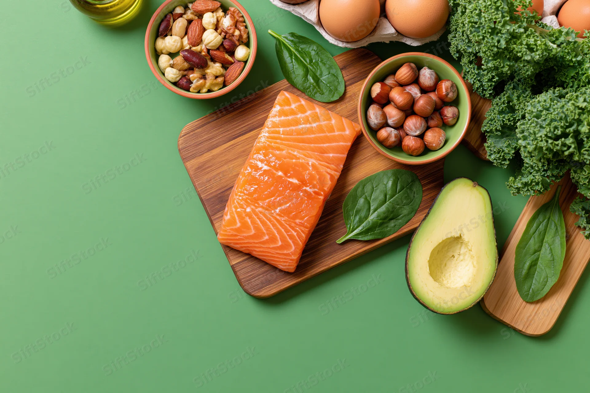 Salmon fillet and avocado on wooden cutting board with nuts, eggs, kale and olive oil arranged on green surface