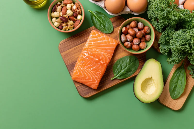 Salmon fillet and avocado on wooden cutting board with nuts, eggs, kale and olive oil arranged on green surface