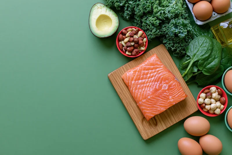 Salmon fillet on wooden cutting board surrounded by keto diet ingredients including eggs, avocado, kale, spinach, and nuts arranged overhead on green surface