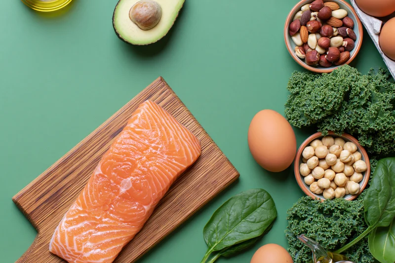 Salmon fillet on a wooden cutting board surrounded by keto diet ingredients including avocado, eggs, nuts, kale and olive oil on a green surface