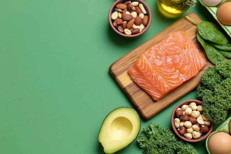 Raw salmon fillet on wooden cutting board surrounded by avocado, kale, nuts, eggs and olive oil on green surface