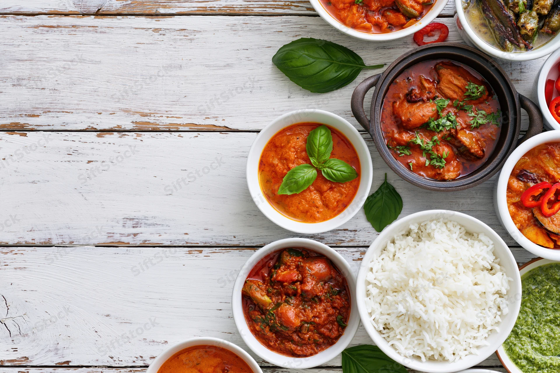 Multiple indian and asian curry dishes arranged on a white wooden table with rice and garnishes in bowls, overhead view