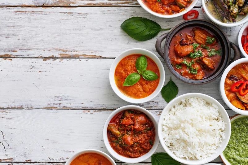 Multiple indian and asian curry dishes arranged on a white wooden table with rice and garnishes in bowls, overhead view
