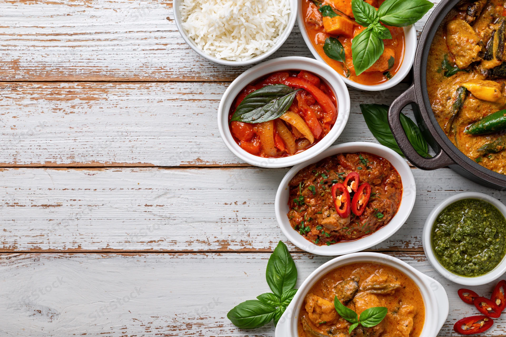 Multiple curry dishes arranged on a white wooden table with bowls of rice and garnishes in a flat lay overhead view
