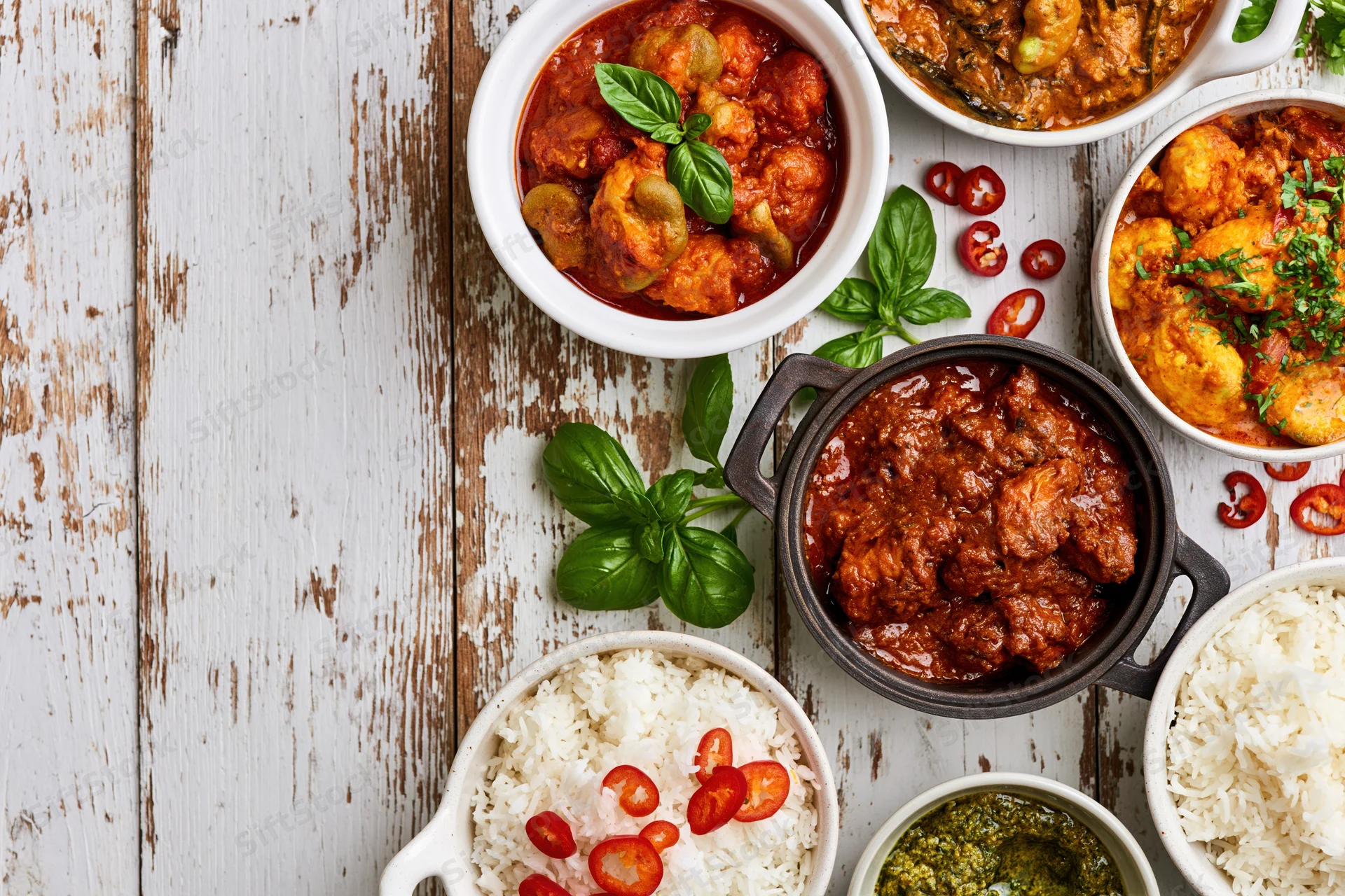 Multiple indian and asian curry dishes with rice and herbs arranged on a rustic wooden table in overhead flat lay view