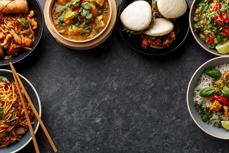 Multiple asian dishes arranged in a flat lay on a dark textured surface with copyspace in center