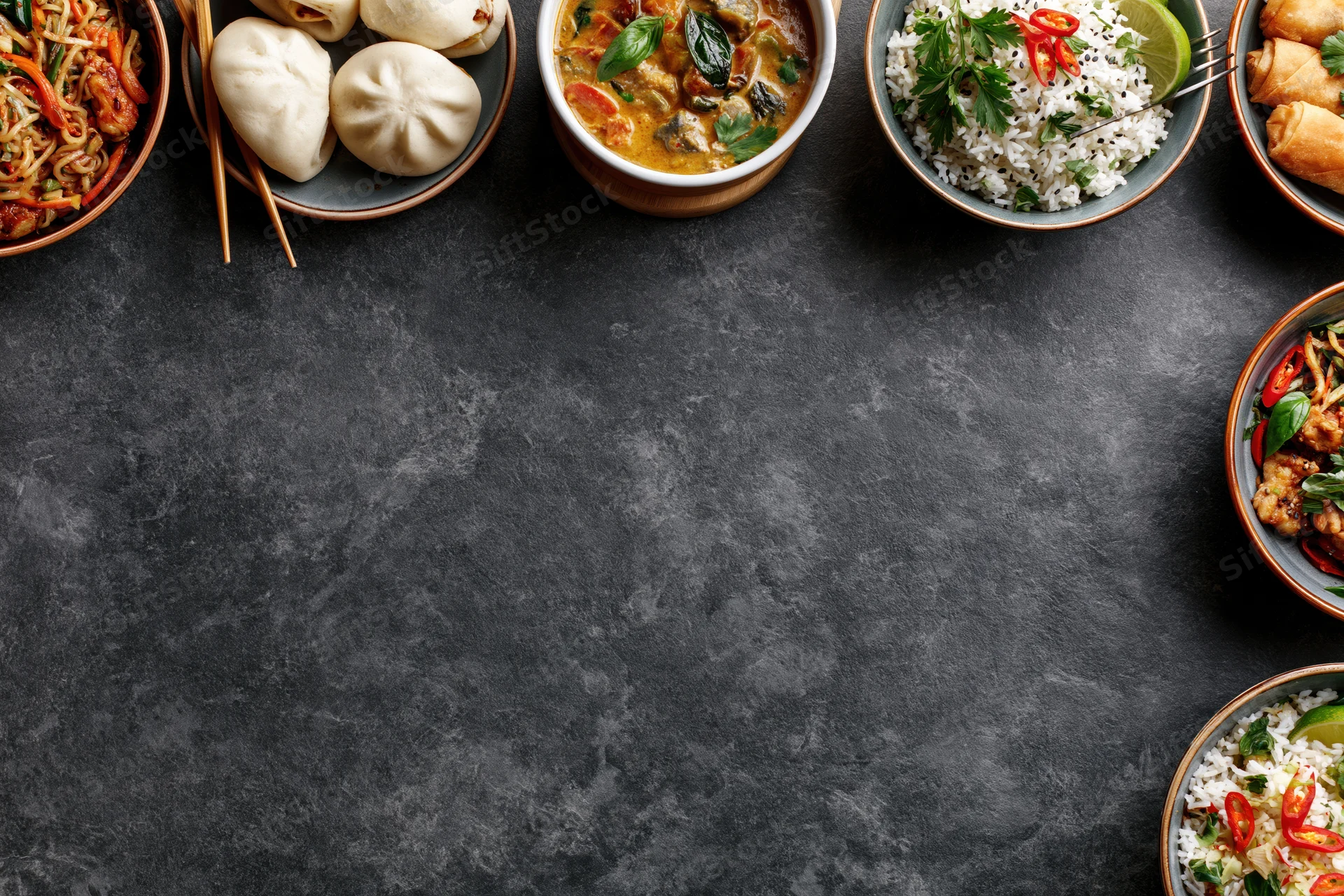 Multiple asian dishes arranged along the edge of a dark textured table in overhead flat lay view with copyspace in center