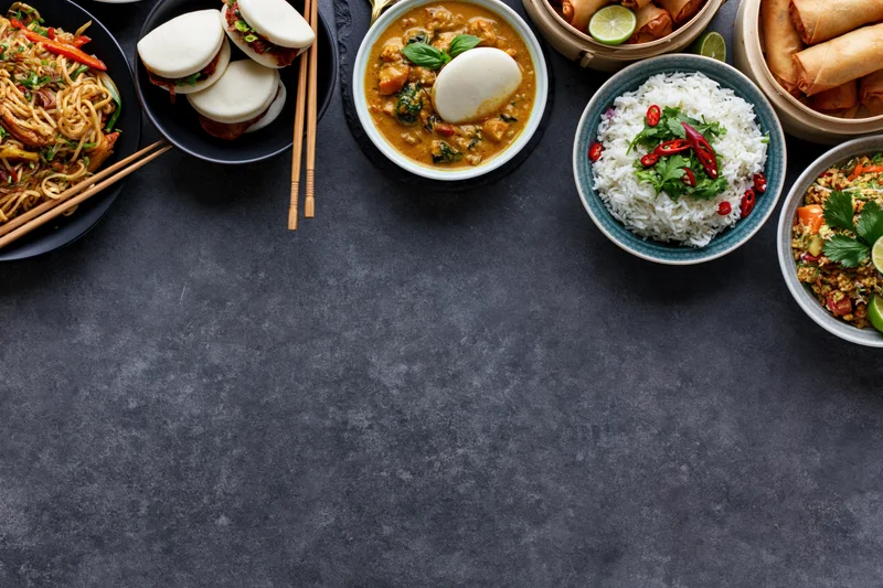 Multiple asian dishes arranged along the top of a dark textured table in overhead flat lay view