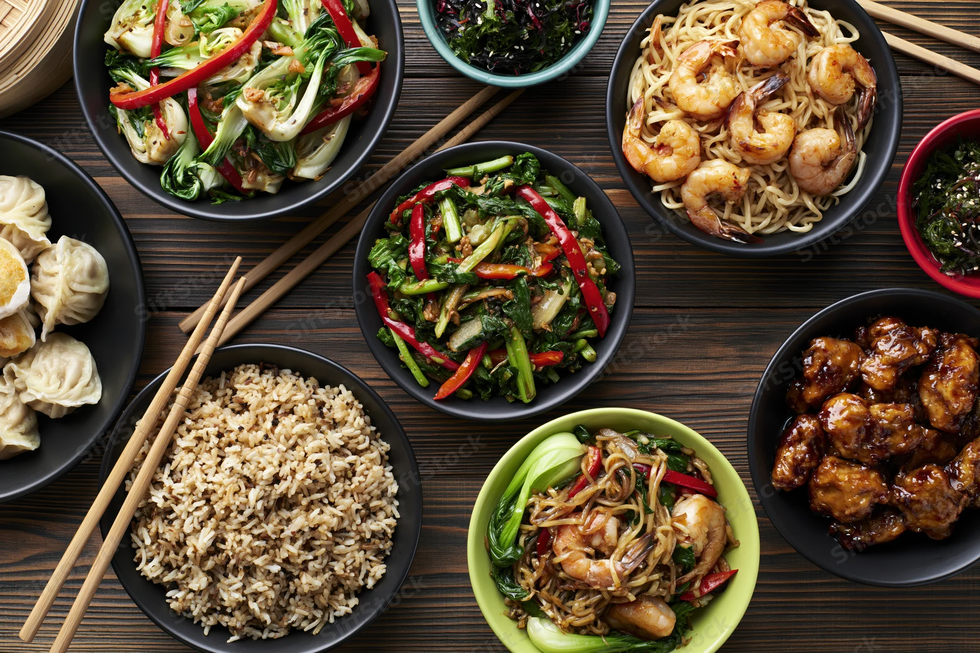 Multiple asian dishes arranged in bowls on a dark wooden table with chopsticks and rice