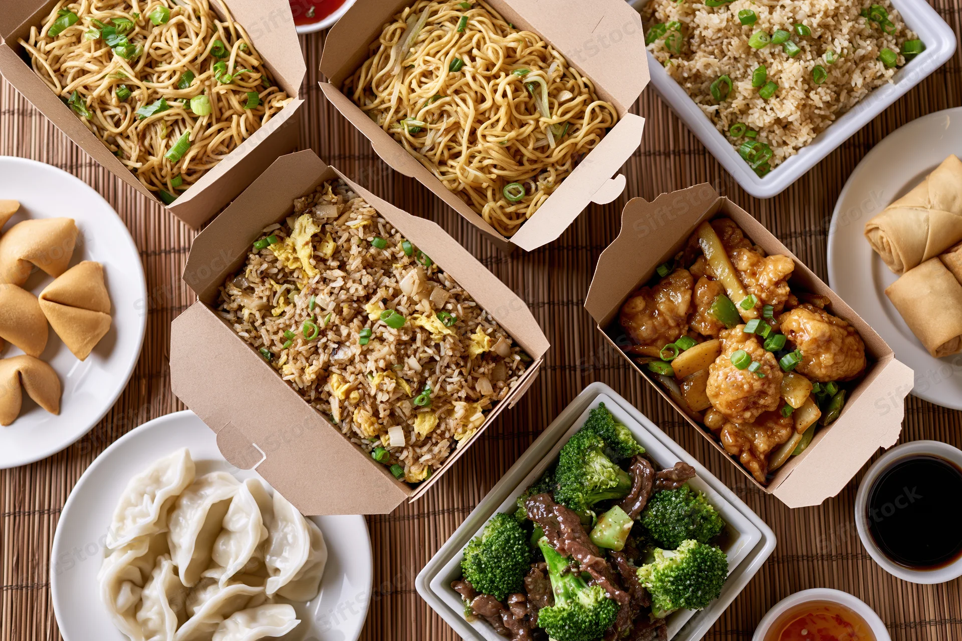 Overhead flat lay of multiple chinese takeout boxes filled with noodles, fried rice, general tso's chicken, broccoli beef, dumplings, wontons, egg rolls, and sauces on a bamboo mat