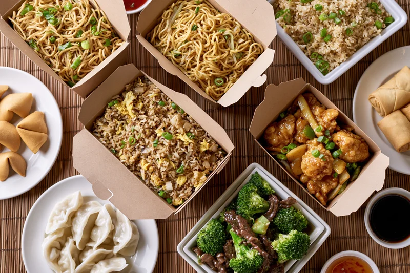Overhead flat lay of multiple chinese takeout boxes filled with noodles, fried rice, general tso's chicken, broccoli beef, dumplings, wontons, egg rolls, and sauces on a bamboo mat
